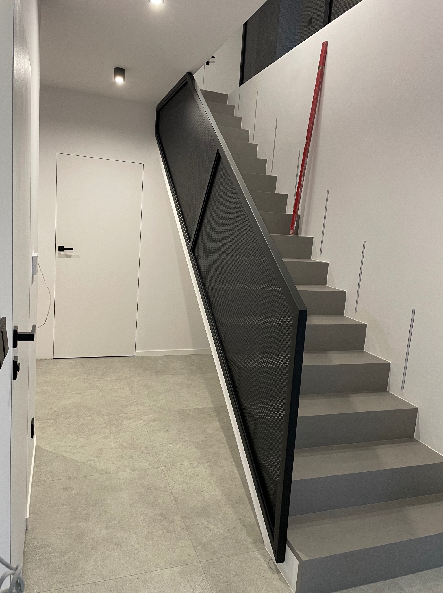 Nowoczesne schody z metalową balustradą w Nowinie. Minimalistyczny design, szare stopnie i czarna siatka balustrady. Widoczny poziomica oparta o ścianę.