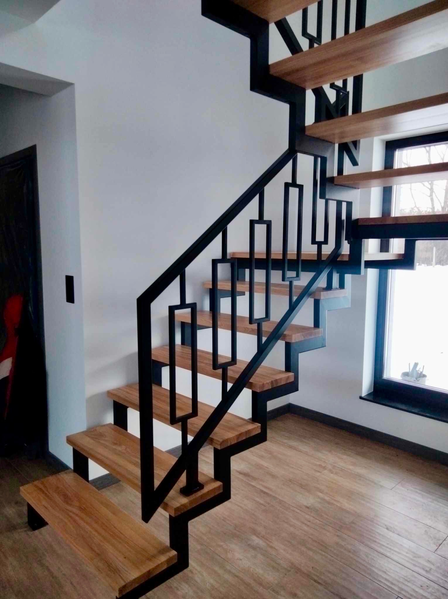 Nowoczesne schody z drewnianymi stopniami i czarną, geometryczną balustradą metalową, widoczne z dołu, z oknem w tle.