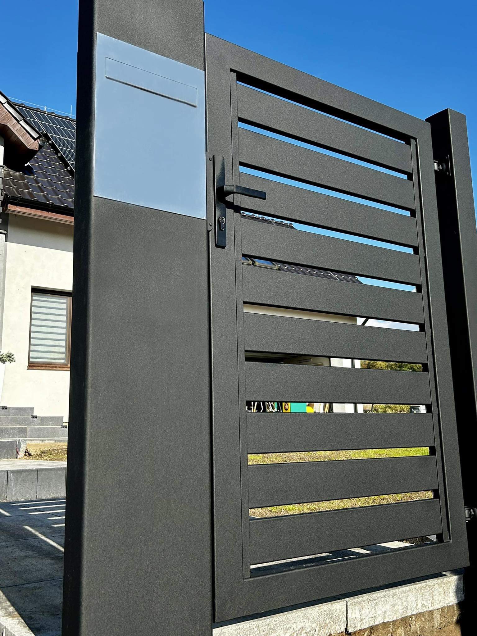 Nowoczesna, grafitowa brama posesyjna z poziomymi szczeblami i skrzynką na listy, na tle domu z panelami słonecznymi i błękitnego nieba.