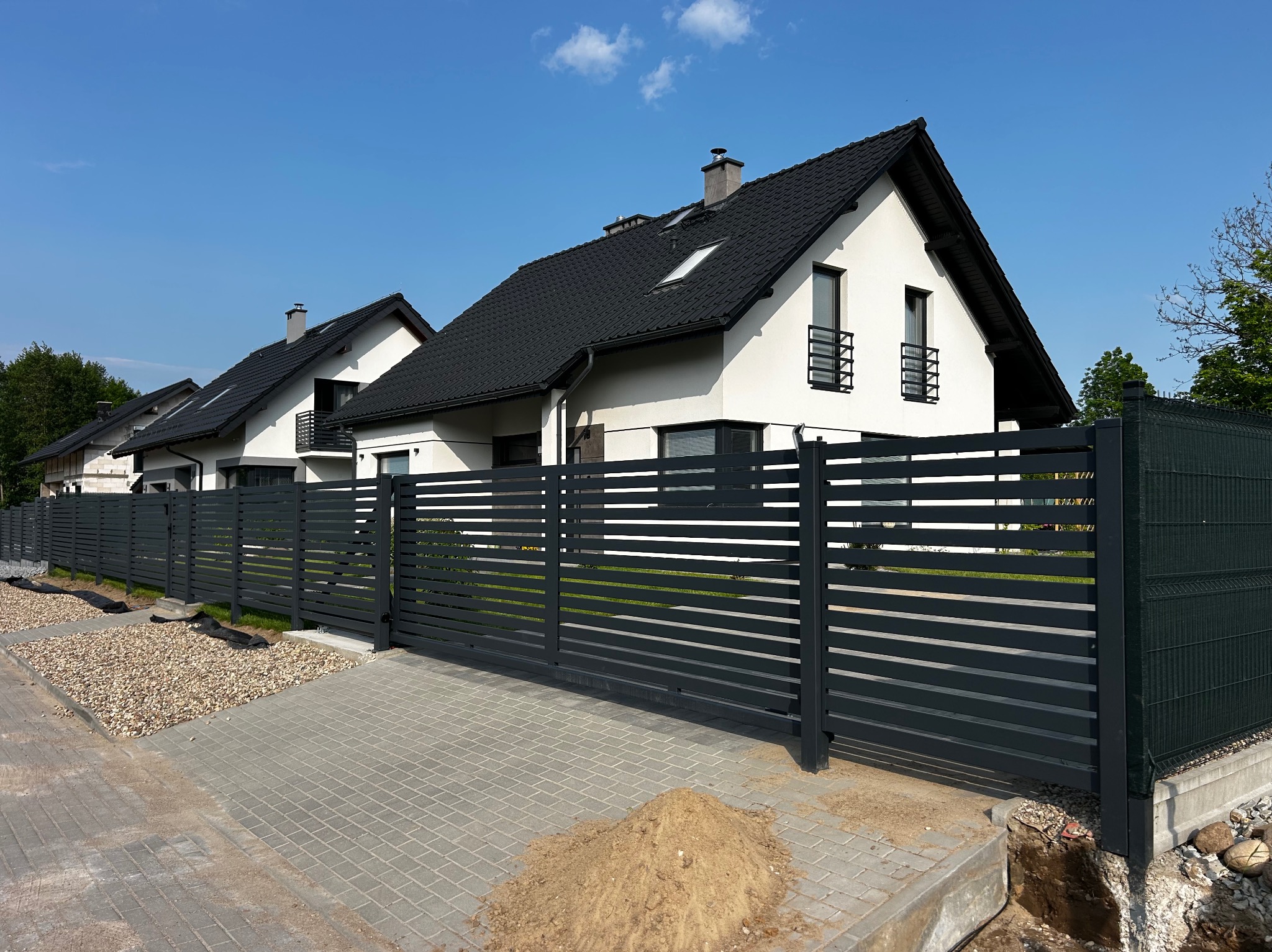 Nowoczesne, poziome ogrodzenie posesji z przesuwną bramą w kolorze grafitowym, na tle białych domów z czarnymi dachami i błękitnego nieba.