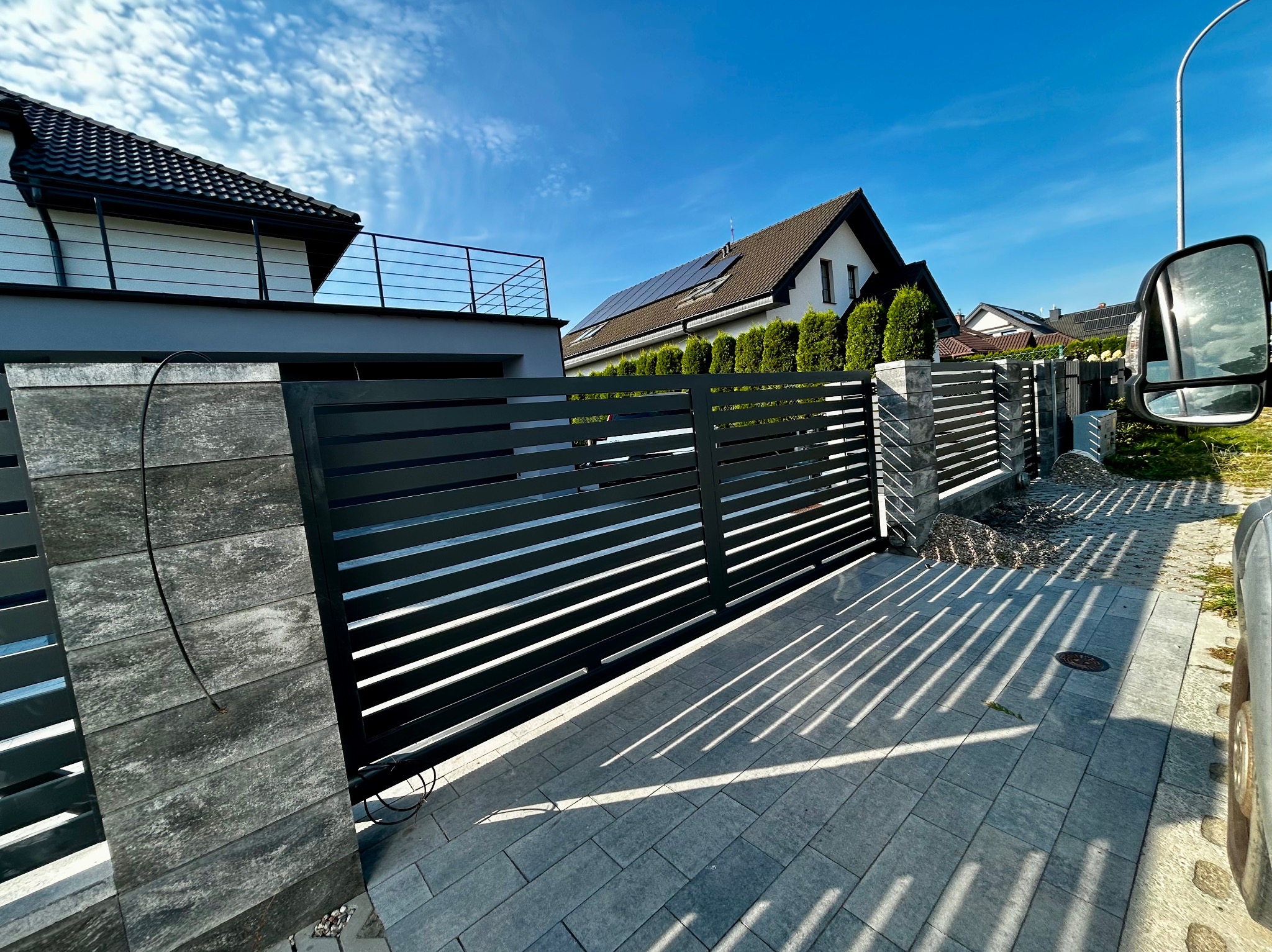 Nowoczesna, pozioma brama wjazdowa z szarymi, betonowymi słupkami, rzucająca cienie na szarą kostkę brukową. W tle domy jednorodzinne i błękitne niebo.