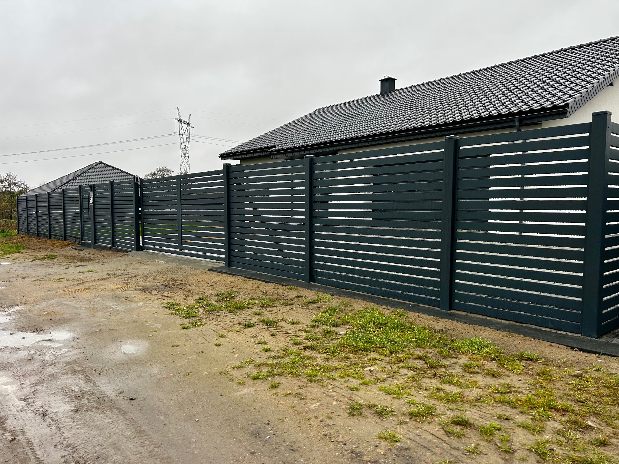 Nowoczesne, poziome ogrodzenie panelowe w kolorze antracytowym, z widoczną bramą wjazdową, na tle domu z ciemną dachówką i pochmurnego nieba.