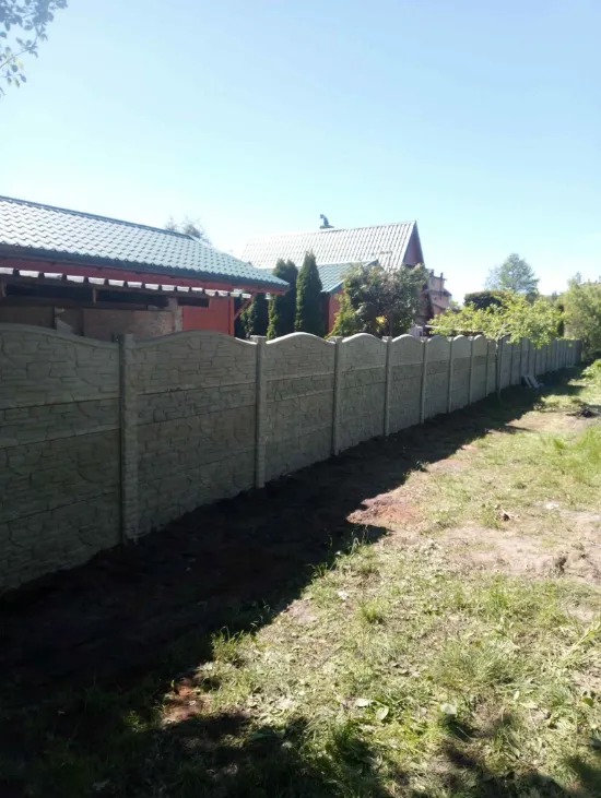 Długi, szary, betonowy płot z wzorem kamienia, oddzielający posesję z zielonym dachem od trawnika. Widok z poziomu gruntu, w tle domy i drzewa.