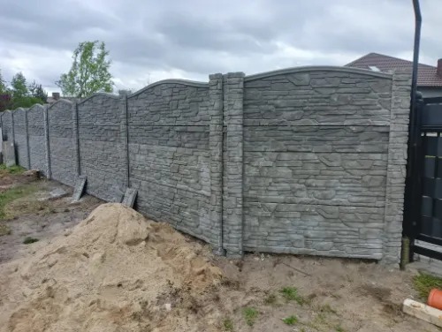 Szary, betonowy płot z imitacją kamienia, z łukowatym wykończeniem, widoczny z perspektywy ziemi, z kupką piasku u podstawy. Fragment bramy w tle.