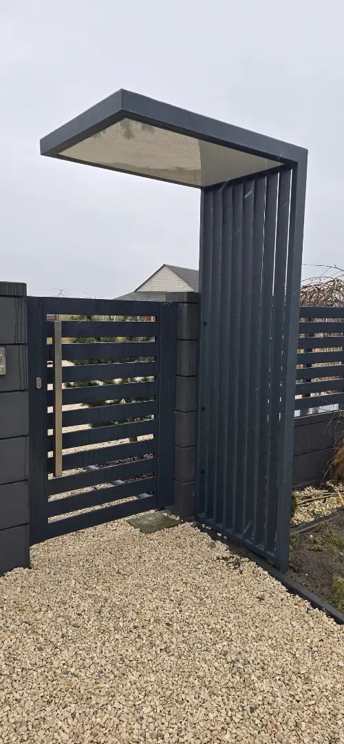 Nowoczesna, grafitowa furtka z poziomymi szczeblami i zadaszeniem, prowadząca na posesję wysypaną jasnym żwirem. Widoczny fragment ogrodzenia z betonowych bloczków.