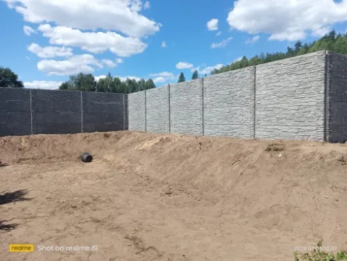 Długi, szary mur z paneli imitujących kamień, otaczający teren z ziemią i małym, czarnym zbiornikiem. Błękitne niebo z chmurami w tle.