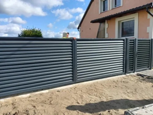 Nowoczesna, pozioma brama wjazdowa z metalowymi lamelami w kolorze grafitowym, z widoczną furtką po prawej stronie, na tle domu jednorodzinnego i błękitnego nieba.