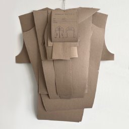GARMENT X STUDIO - Kreatywna kompozycja z papierowych wykrojów odzieży, zawieszona na ścianie, z etykietą Garment X Studio. Widoczne detale kroju kurtki.