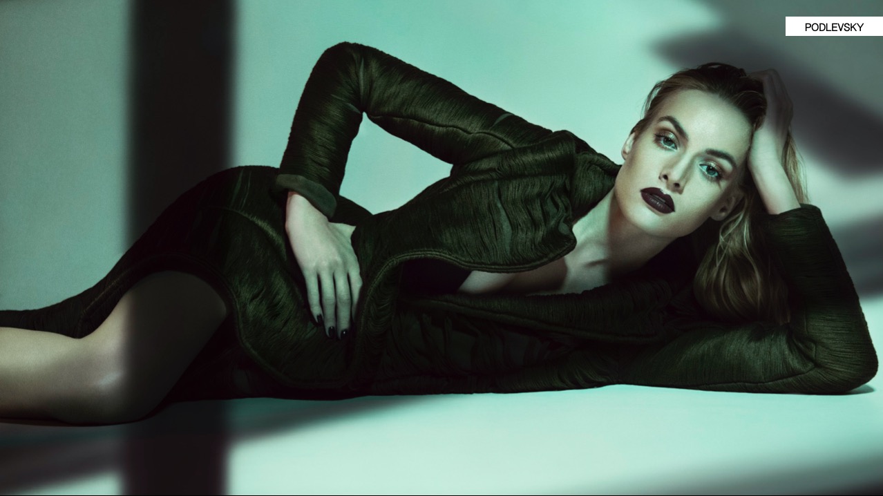 Leżąca modelka w ciemnej, teksturowanej sukience i marynarce, z mocnym makijażem ust, w chłodnym oświetleniu studyjnym. Ujęcie z boku, podkreślające fakturę materiału.