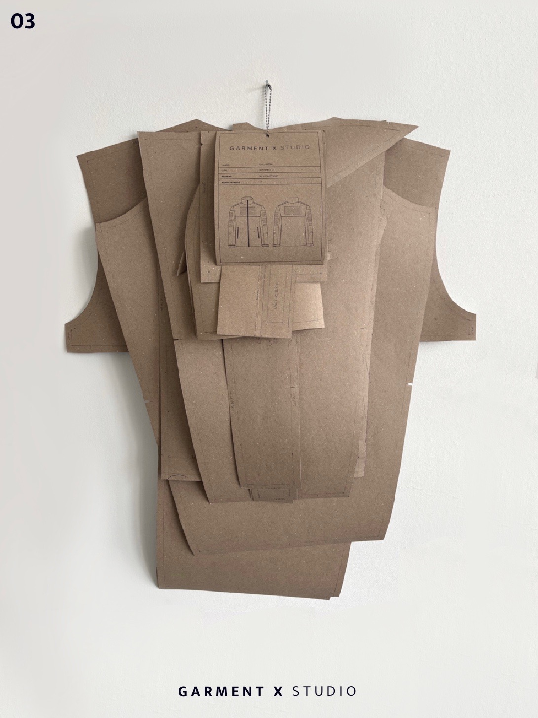 Kreatywna kompozycja z papierowych wykrojów odzieży, zawieszona na ścianie, z etykietą Garment X Studio. Widoczne detale kroju kurtki.