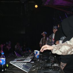 SZTUKA NATALIA WAWER - DJ na scenie klubowej, miksujący muzykę dla publiczności. Widoczny sprzęt DJ-ski, laptopy, napoje i tłum ludzi w tle. Scena oświetlona kolorowymi światłami.