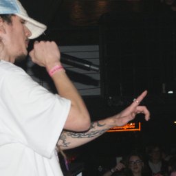 SZTUKA NATALIA WAWER - Młody mężczyzna w białej koszulce i czapce rapera, z tatuażami na ramieniu, śpiewa do mikrofonu na scenie podczas koncertu. W tle widoczny tłum.