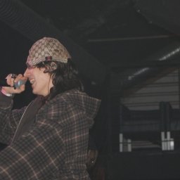 SZTUKA NATALIA WAWER - Wokalista w czapce Gucci, z mikrofonem na scenie, w trakcie występu. Uśmiechnięty, w koszuli w kratę. W tle ciemna scena z elementami oświetlenia.