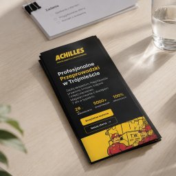 56CREATIVES - Czarna, składana ulotka firmy 'ACHILLES Przeprowadzki' z żółtymi akcentami i ilustracją uśmiechniętych pracowników, leżąca na jasnym drewnianym biurku obok przybornika i szklanki wody.