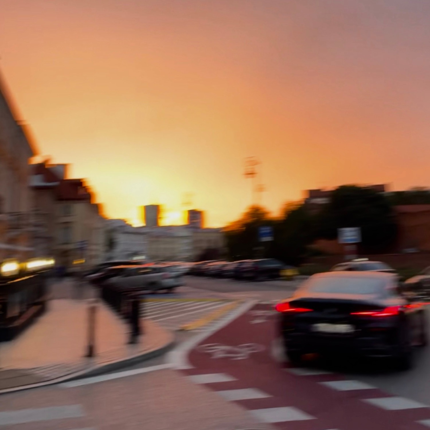 Rozmyte ujęcie ulicy w Warszawie o zmierzchu, z widocznymi budynkami i samochodami na przejściu dla pieszych, w ciepłej tonacji kolorystycznej.