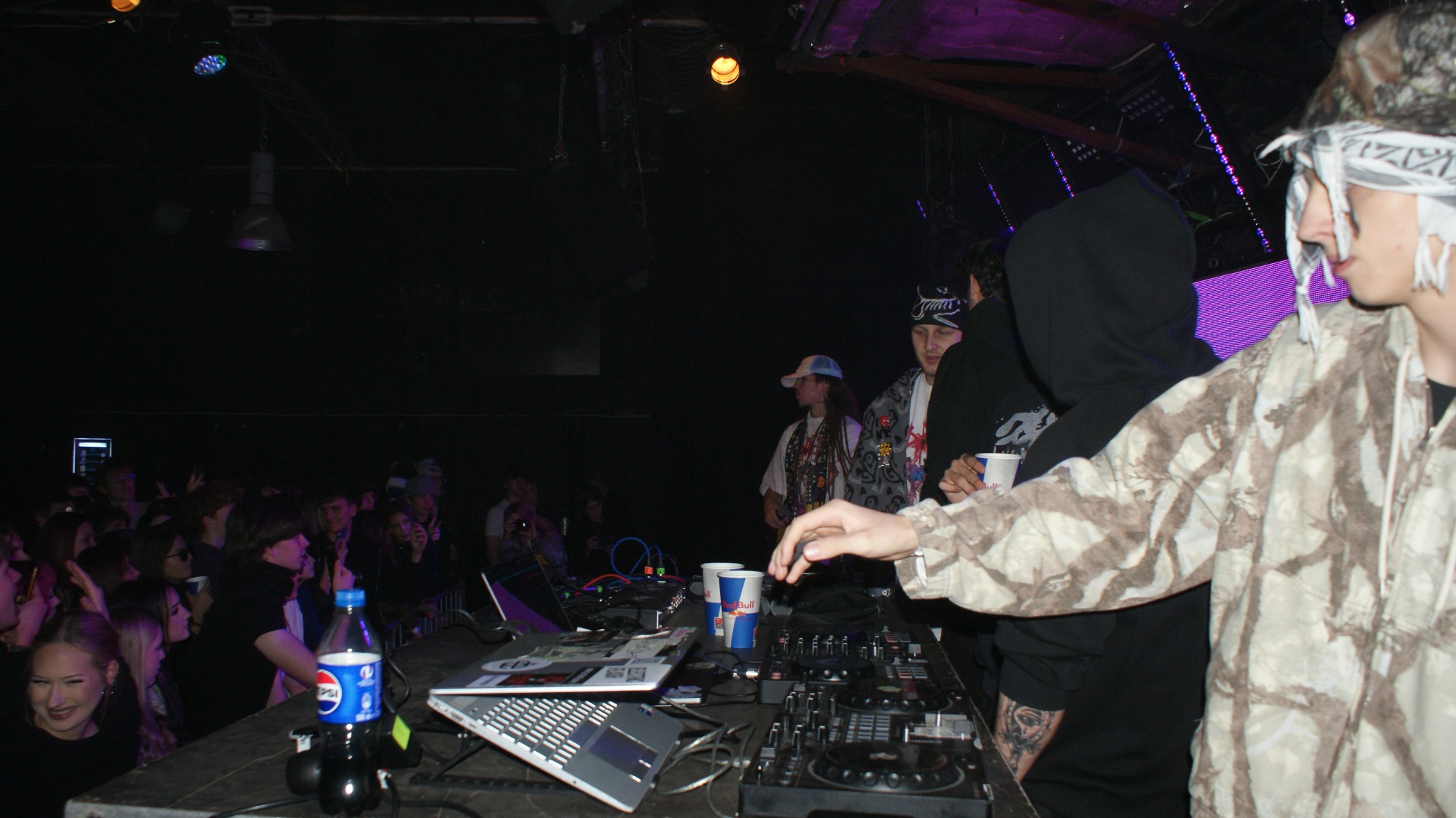 DJ na scenie klubowej, miksujący muzykę dla publiczności. Widoczny sprzęt DJ-ski, laptopy, napoje i tłum ludzi w tle. Scena oświetlona kolorowymi światłami.