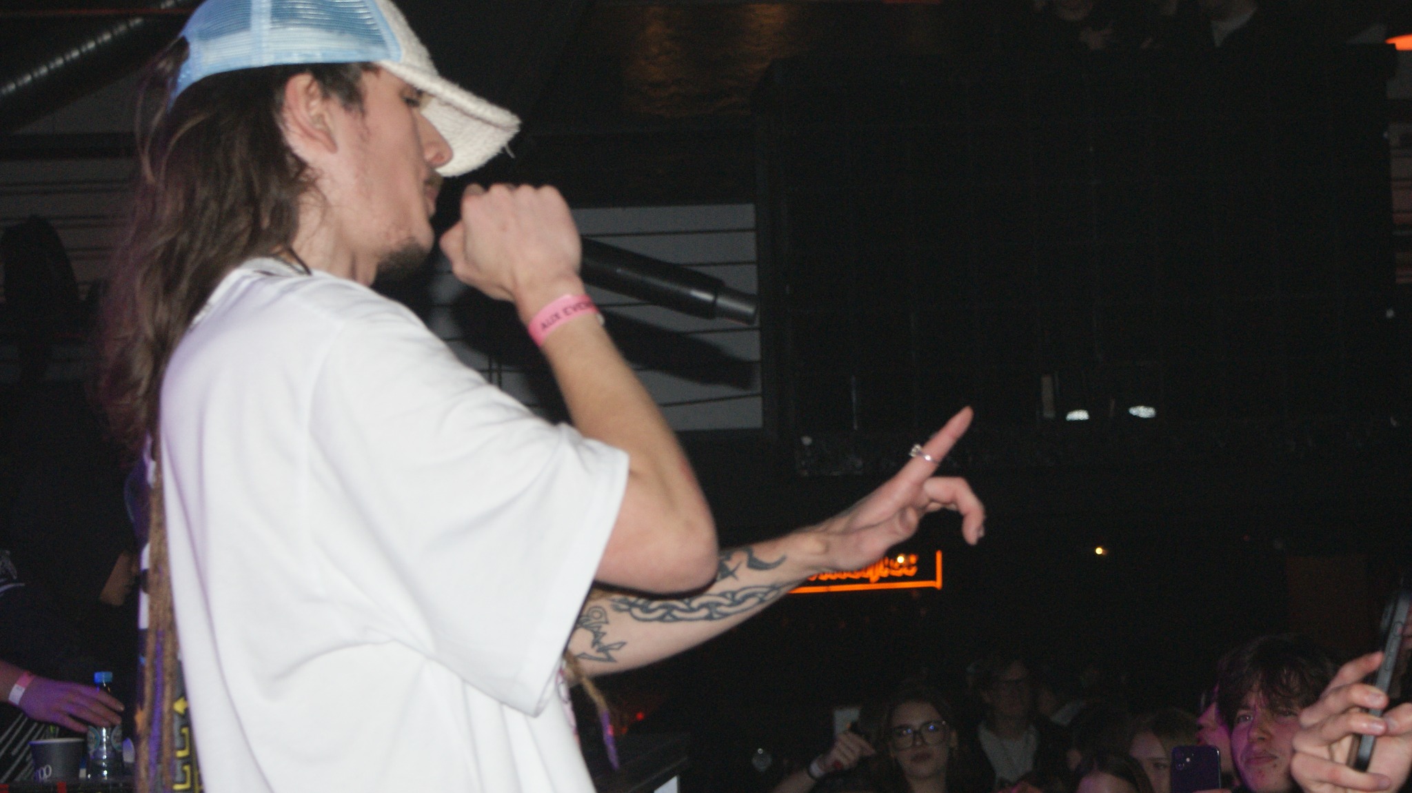Młody mężczyzna w białej koszulce i czapce rapera, z tatuażami na ramieniu, śpiewa do mikrofonu na scenie podczas koncertu. W tle widoczny tłum.
