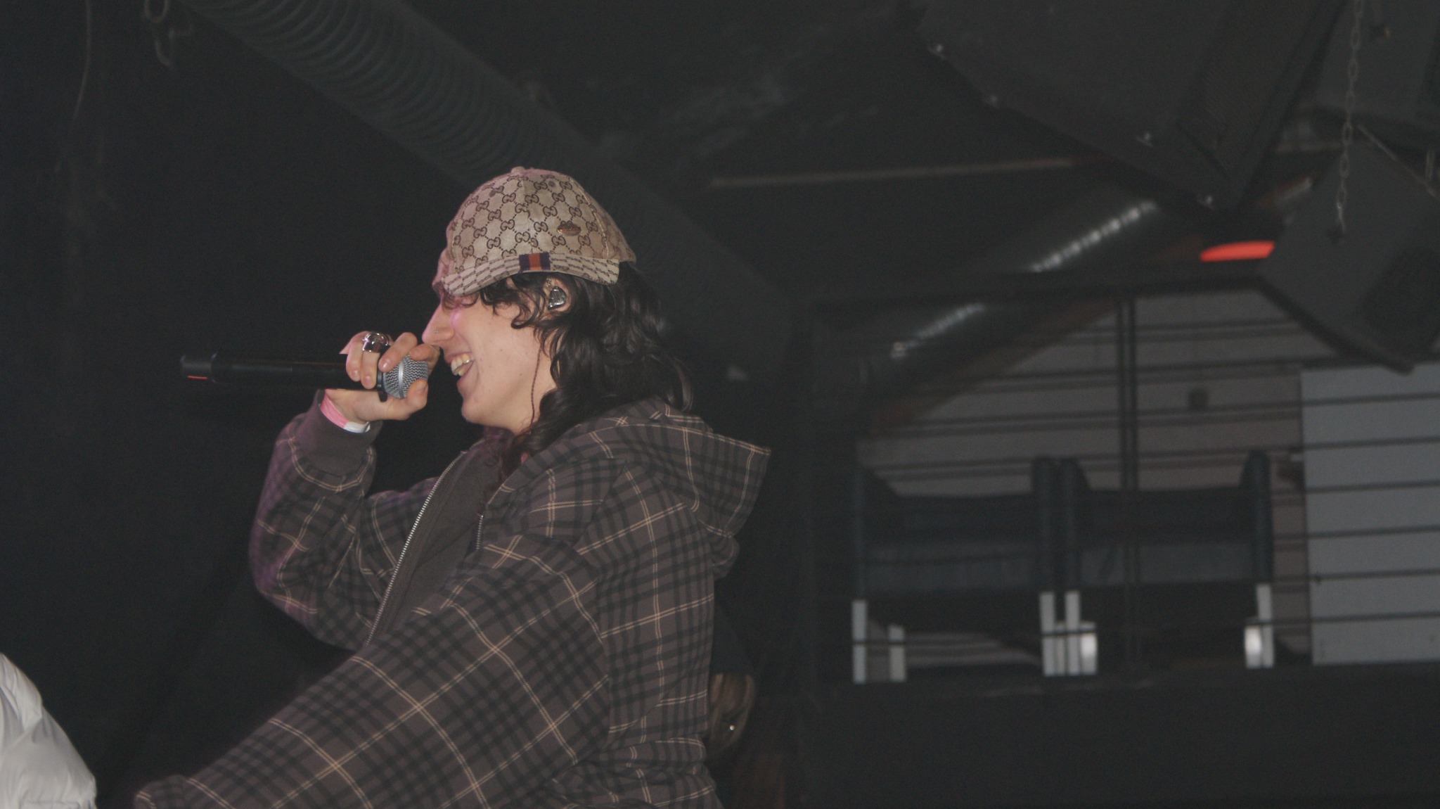 Wokalista w czapce Gucci, z mikrofonem na scenie, w trakcie występu. Uśmiechnięty, w koszuli w kratę. W tle ciemna scena z elementami oświetlenia.