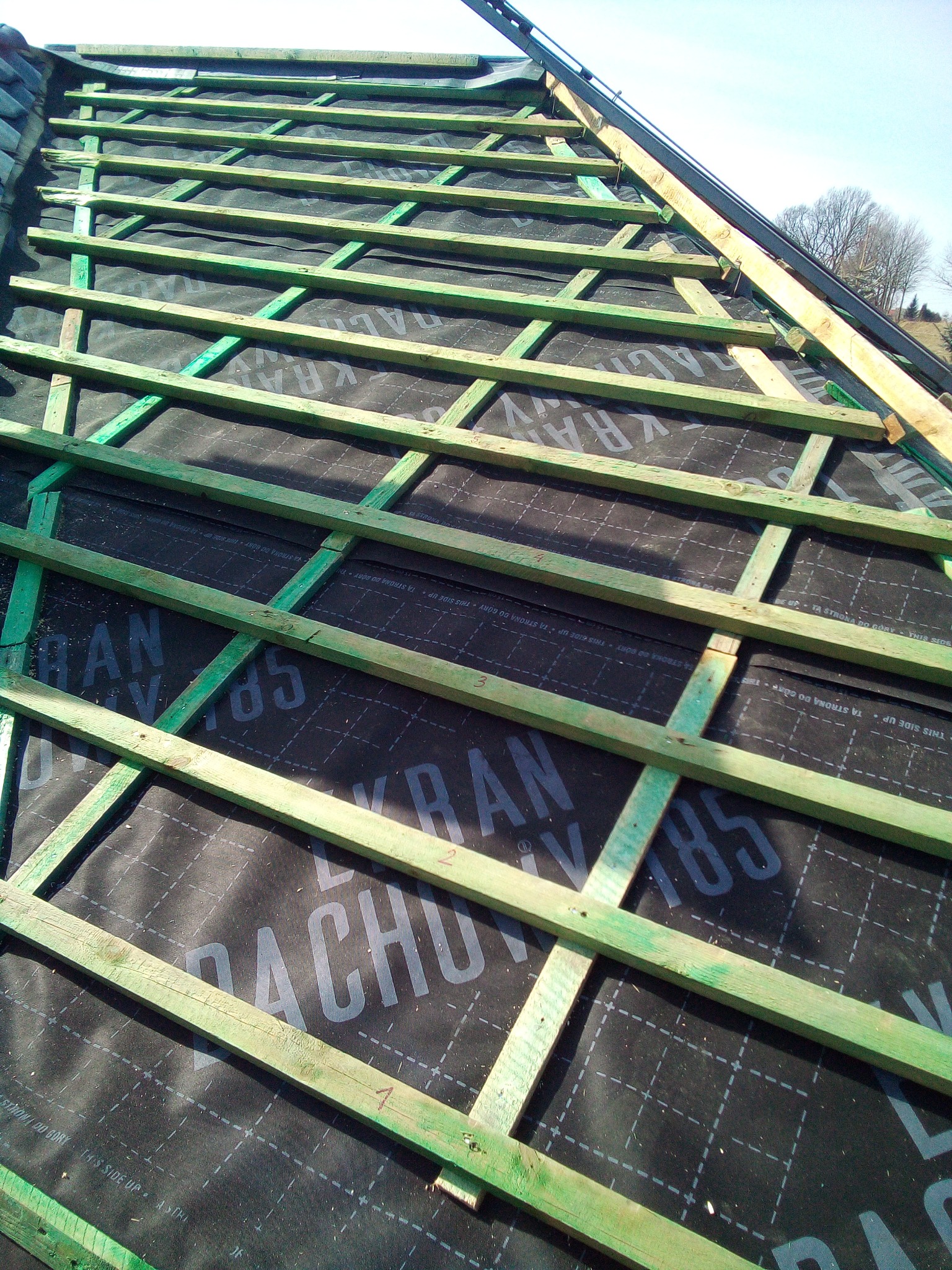 Konstrukcja dachu z zielonych łat na czarnej membranie dachowej z nadrukiem, widok z góry pod kątem. Elementy mocowania widoczne w tle.