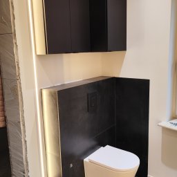 GLAZURKARZ Mateusz Moskwa - Nowoczesna łazienka po remoncie: podwieszana toaleta, czarne płytki, szafki i oświetlenie LED. Widoczny fragment niedokończonego wejścia do łazienki.