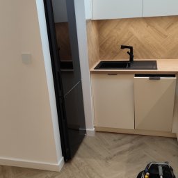 GLAZURKARZ Mateusz Moskwa - Nowoczesna kuchnia z czarną lodówką Samsung, drewnianym blatem i wzorem jodełki na ścianie. Na podłodze panele w jodełkę. Widoczna torba z narzędziami.