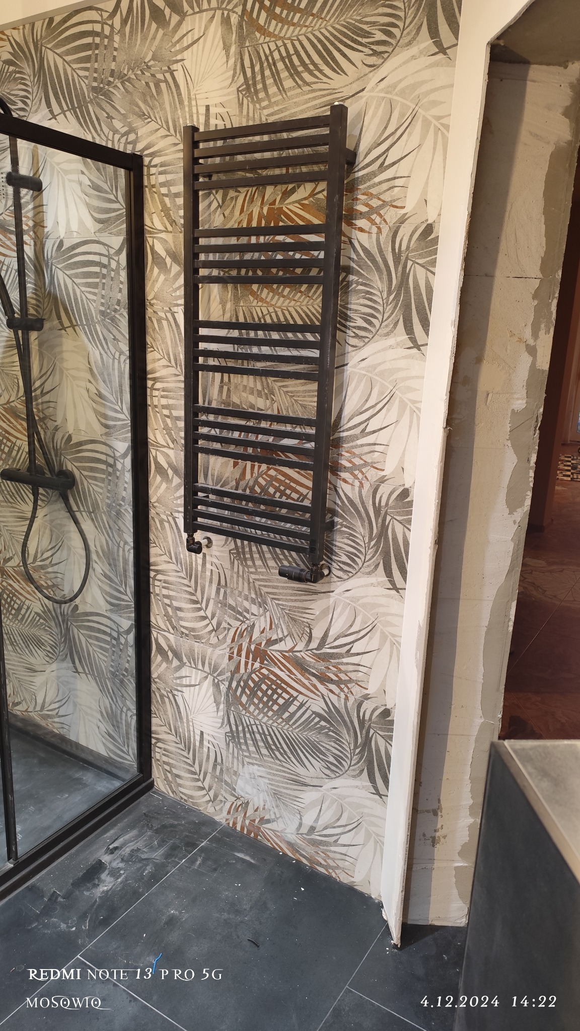 Nowoczesna łazienka z czarną kabiną prysznicową, grzejnikiem drabinkowym na ścianie w liście i ciemną podłogą. Widoczny fragment futryny drzwi.