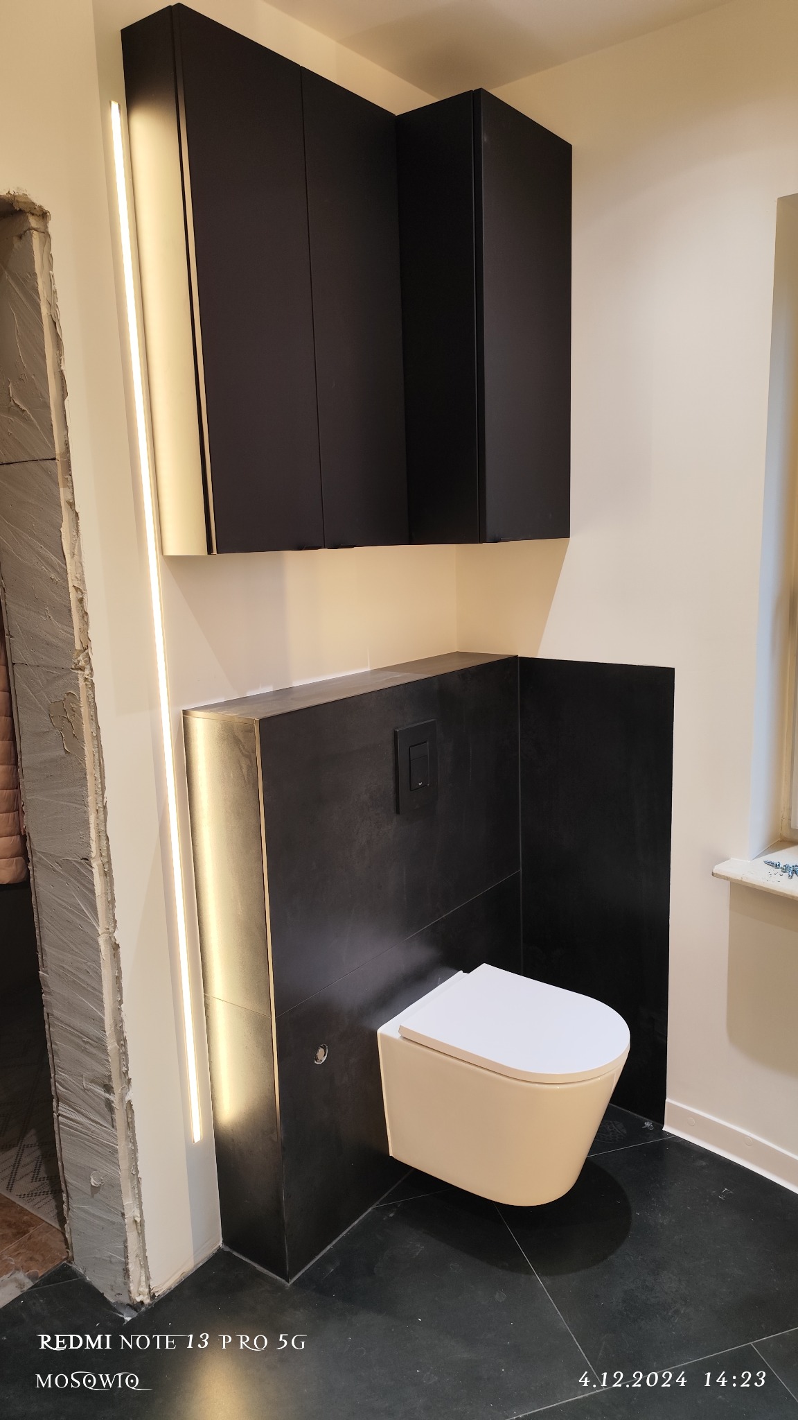 Nowoczesna łazienka po remoncie: podwieszana toaleta, czarne płytki, szafki i oświetlenie LED. Widoczny fragment niedokończonego wejścia do łazienki.