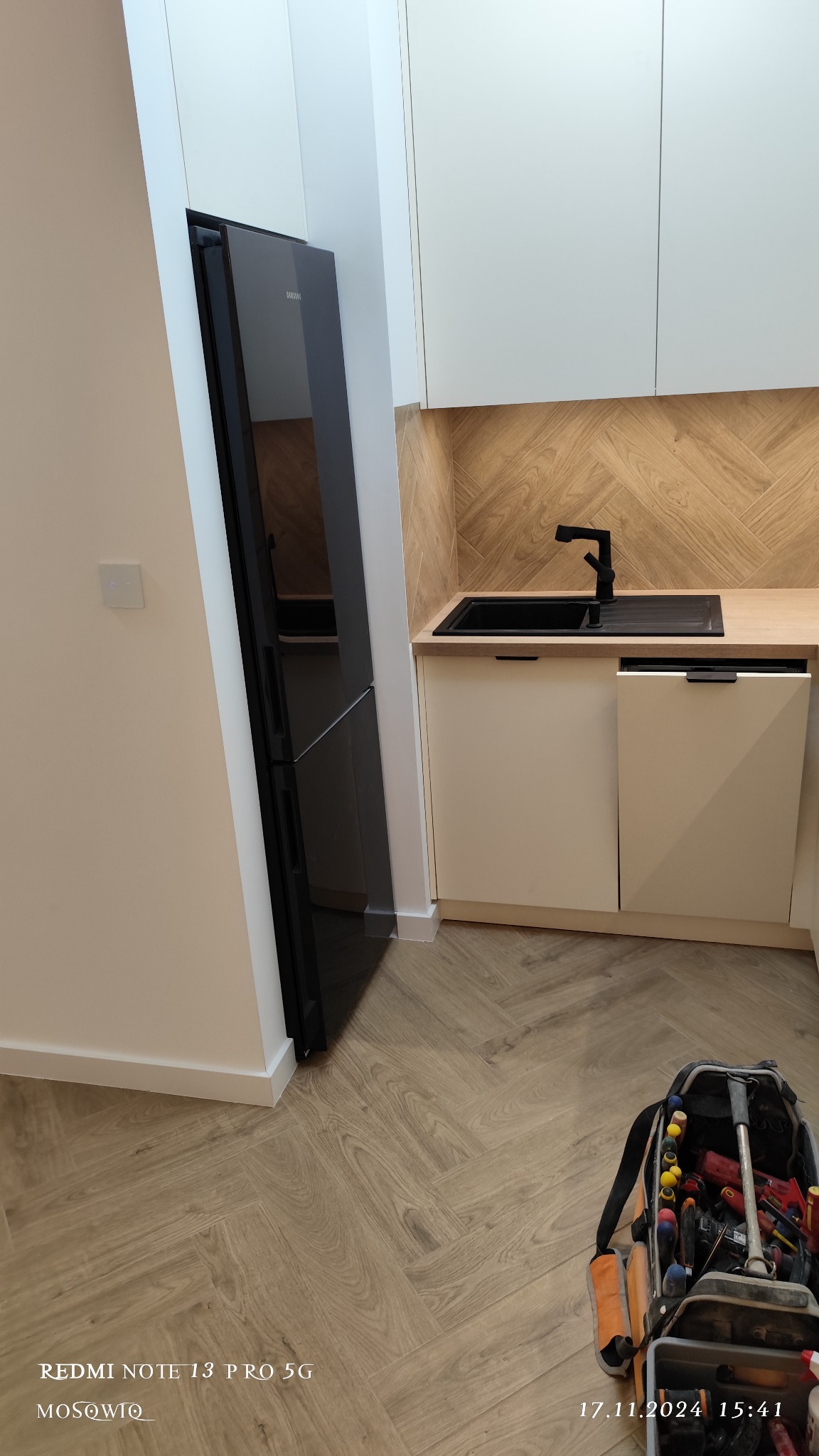 Nowoczesna kuchnia z czarną lodówką Samsung, drewnianym blatem i wzorem jodełki na ścianie. Na podłodze panele w jodełkę. Widoczna torba z narzędziami.