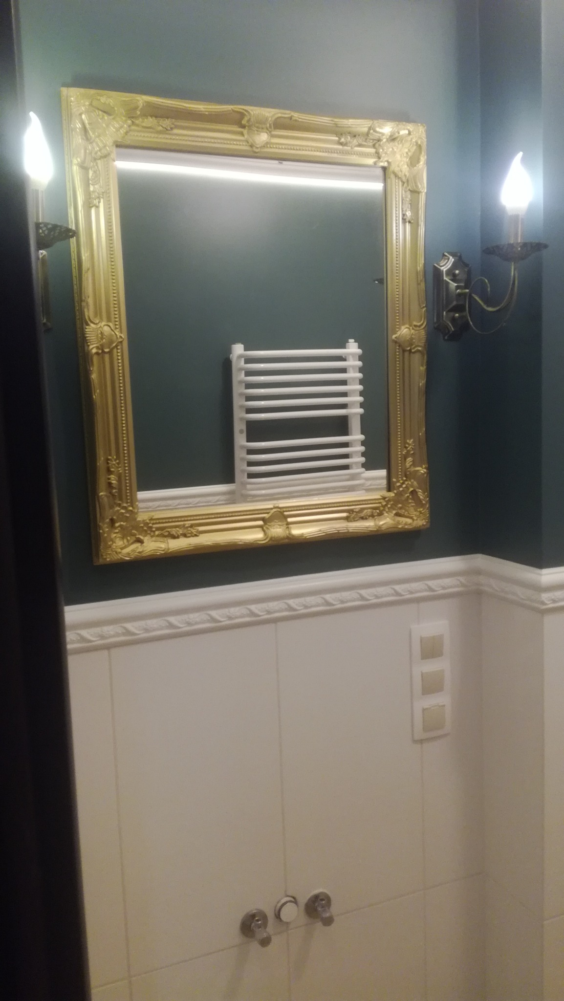 Lustro w złotej, zdobionej ramie odbija biały grzejnik drabinkowy na tle ciemnozielonej ściany w wykończonej łazience z białymi płytkami i dekoracyjnymi kinkietami.