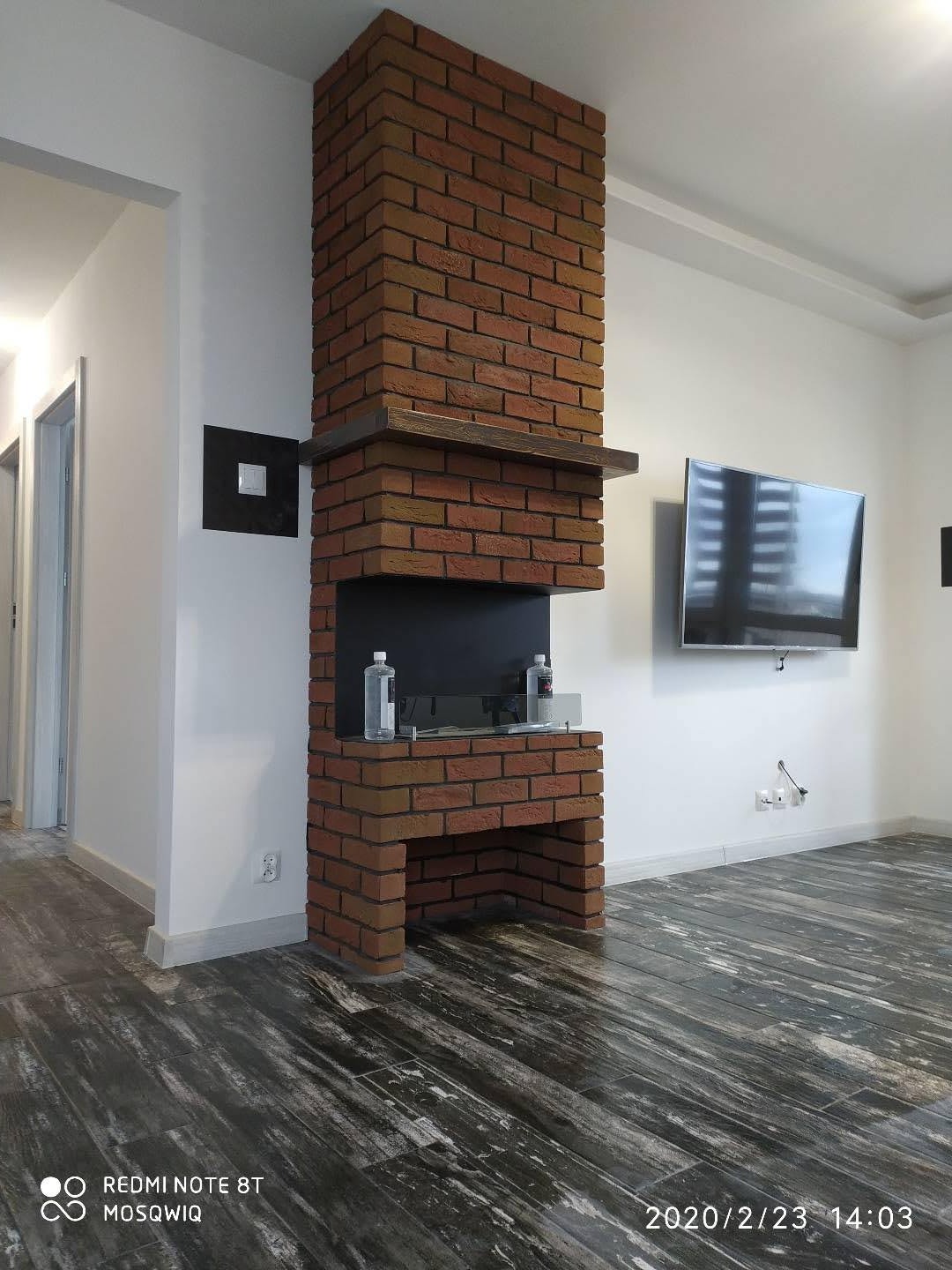 Murowany kominek z cegły klinkierowej w nowoczesnym salonie. Widoczne ciemne, drewnopodobne płytki podłogowe i telewizor na ścianie. Minimalistyczny design.