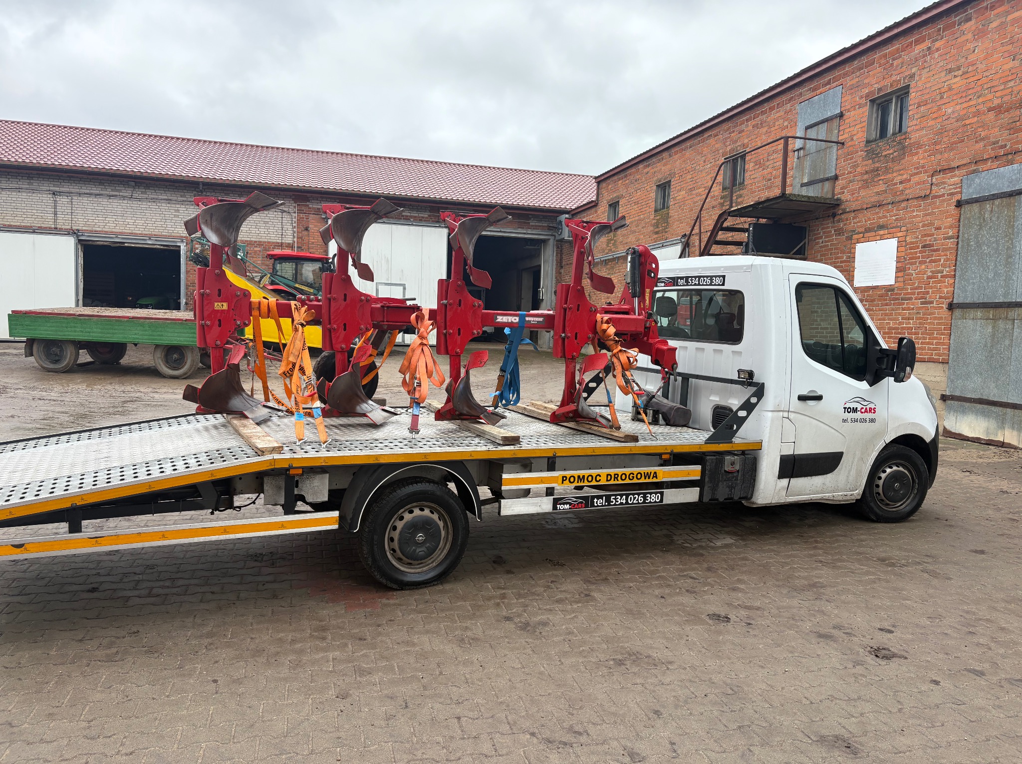 Laweta z czerwonym pługiem rolniczym Zeto System, zabezpieczonym pomarańczowymi pasami. Biały bus z napisem 'Pomoc Drogowa' i logo firmy Tom-Cars na tle ceglanego budynku.