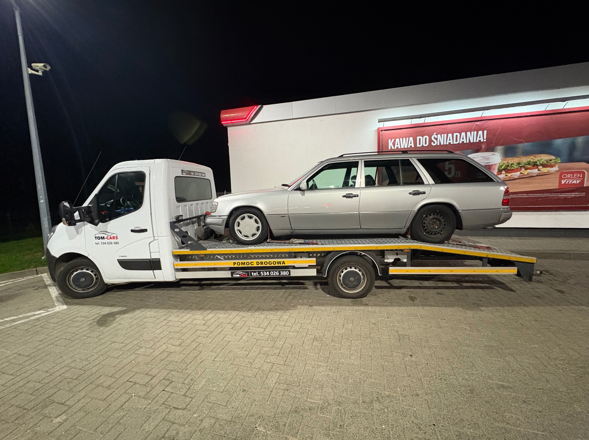 Srebrne kombi Mercedes na żółtej lawecie pomocy drogowej Tom-Cars, na tle stacji benzynowej Orlen z reklamą kawy i kanapek. Nocne ujęcie.