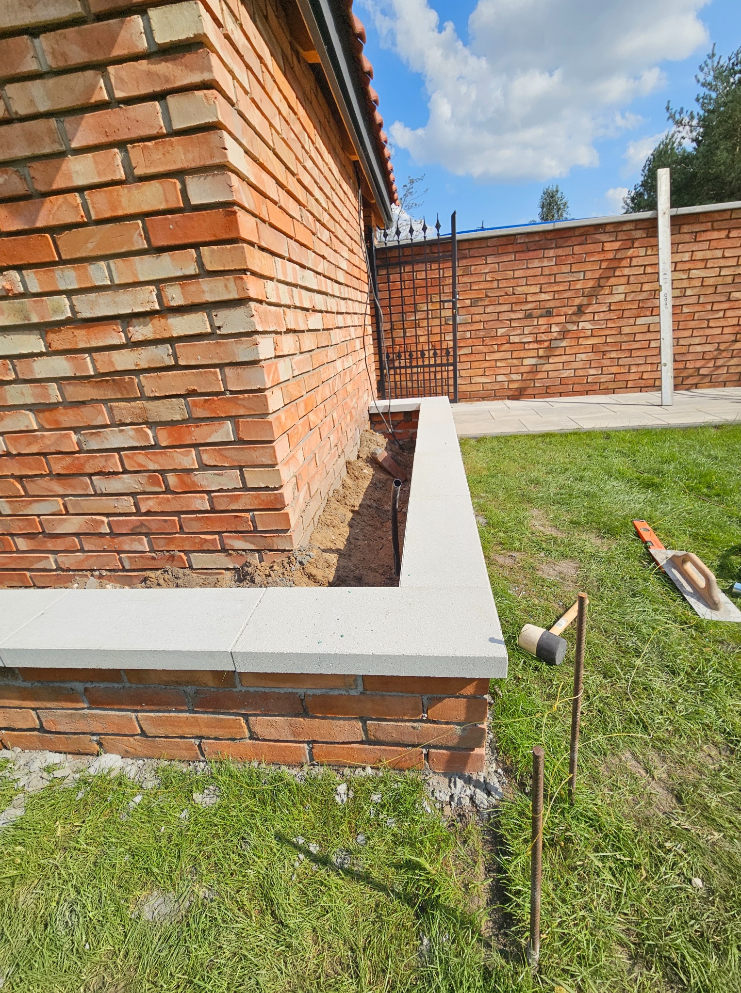 Murowana obudowa rabaty z jasnymi obrzeżami, widoczne narzędzia i fragment trawnika. Elementy ogrodowe w trakcie budowy, ceglana ściana budynku w tle.