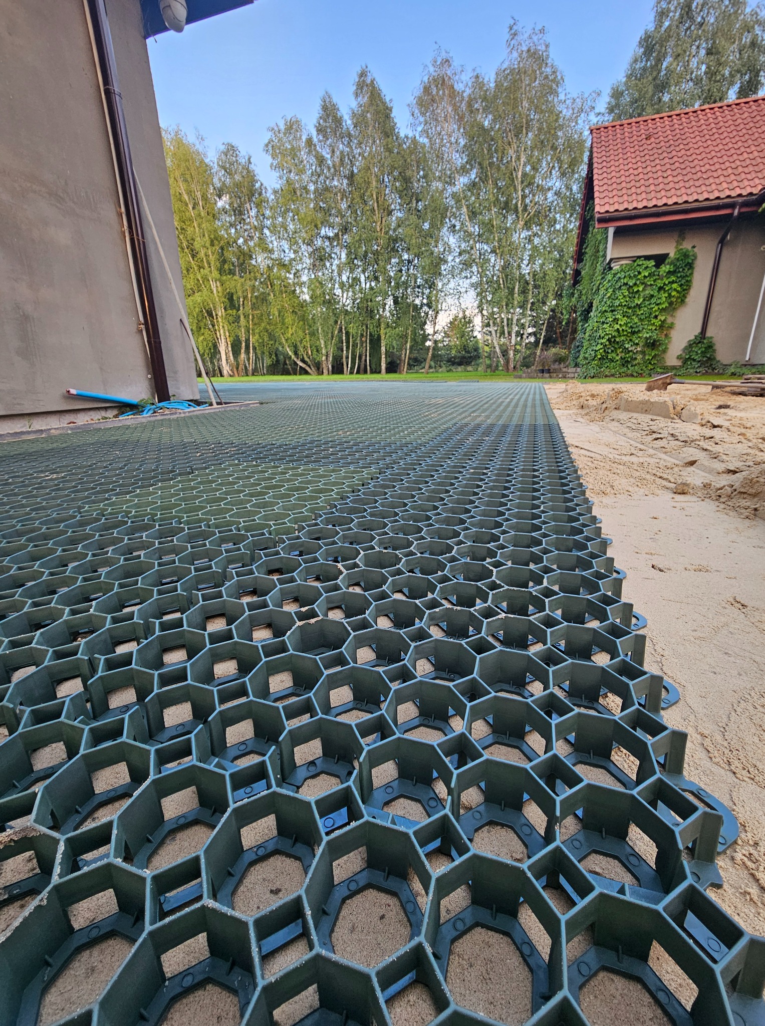 Podjazd z ażurowych płyt z tworzywa wypełnionych piaskiem, widok z bliska. W tle budynek i drzewa. Realizacja w ogrodzie.