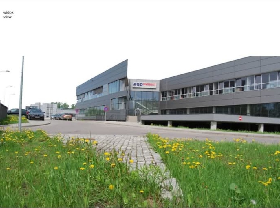 Nowoczesny budynek usługowy AGD Market w Zabrzu z szarą elewacją i dużymi oknami, otoczony zielenią i parkingiem. Widok z poziomu ulicy, wiosenne kwiaty na pierwszym planie.
