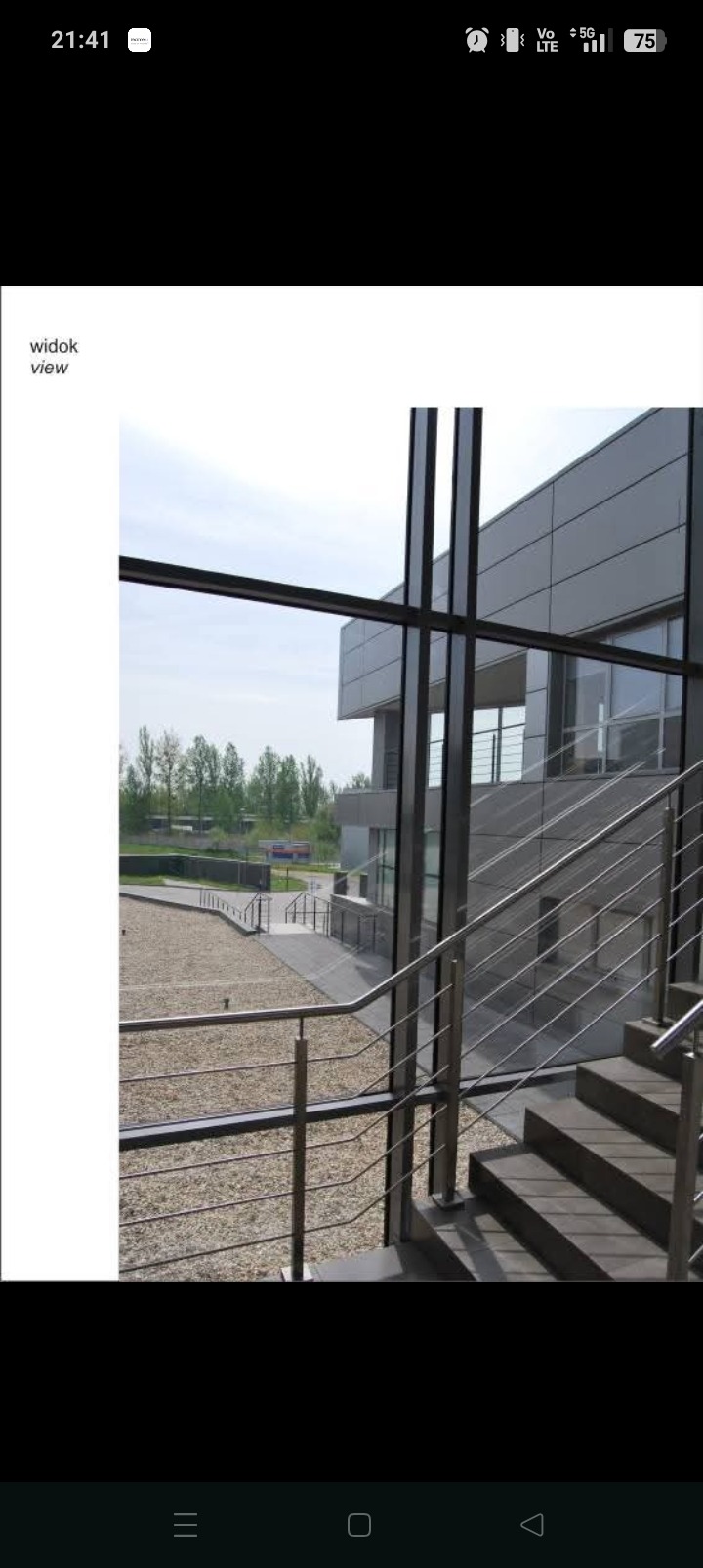 Nowoczesny budynek biurowy z metalowymi schodami i balustradami, widok z wnętrza przez okno na zieleń. Minimalistyczny design, neutralna kolorystyka.