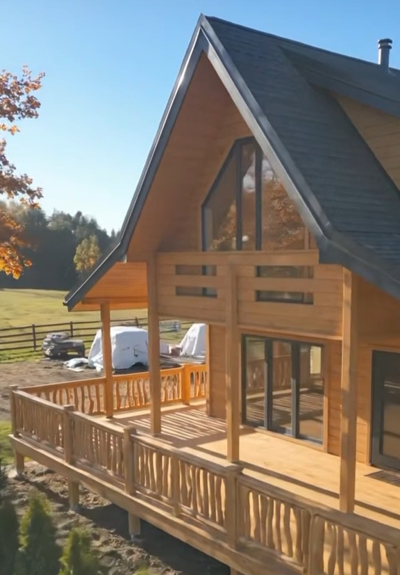 Drewniany dom z balkonem w stylu góralskim, z dużymi oknami i charakterystyczną balustradą. W tle zielone drzewa i błękitne niebo.