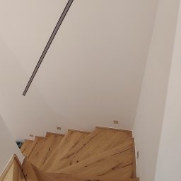 Stanislaw Urbanek - Drewniane schody z widocznymi słojami drewna, oświetlone listwą LED na suficie. Ujęcie z góry podkreśla spiralny kształt schodów i naturalną teksturę drewna.
