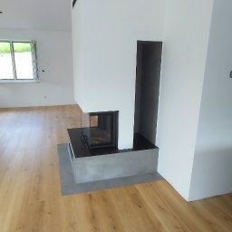 Stanislaw Urbanek - Nowoczesny kominek z szarą obudową w jasnym wnętrzu. Parkiet z naturalnego drewna, białe ściany i okno w tle. Minimalistyczny design.