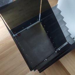 Stanislaw Urbanek - Nowoczesna balustrada schodowa z czarnego szkła, zamontowana na drewnianych schodach. Widoczne detale mocowań i odbicia światła. Ujęcie z góry, z widocznymi butami montera.