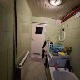 Stanislaw Urbanek - Wnętrze łazienki z zielonymi płytkami, białą wanną i pralką. Widoczne rury, grzejnik i sufit z boazerią. Ściana z ubytkiem tynku. Ujęcie z dołu, perspektywa zniekształcona.