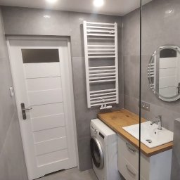 Stanislaw Urbanek - Wykończona łazienka w szarościach z drewnianym blatem, białą armaturą i grzejnikiem drabinkowym. Widoczna pralka i okrągłe lustro z podświetleniem.