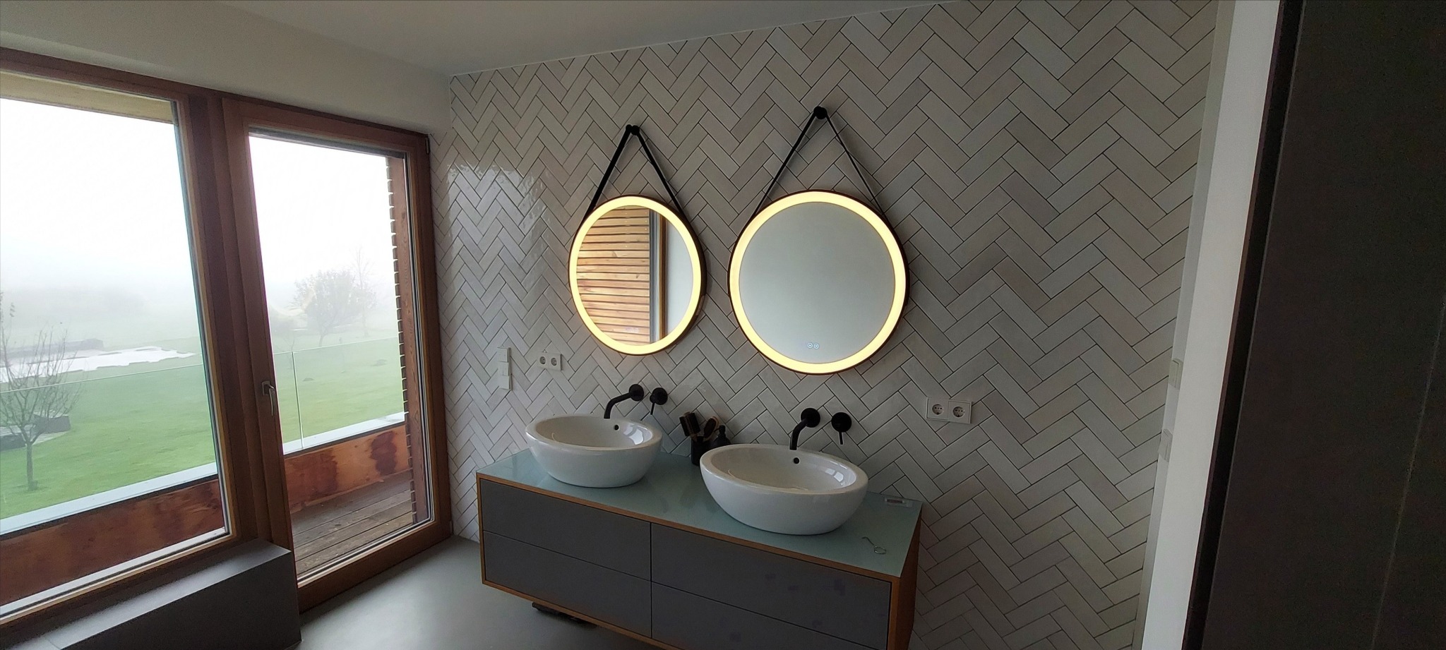 Nowoczesna łazienka z dwoma okrągłymi lustrami z podświetleniem LED, białą armaturą i szarą szafką pod umywalką. Na ścianie białe płytki ułożone w jodełkę.