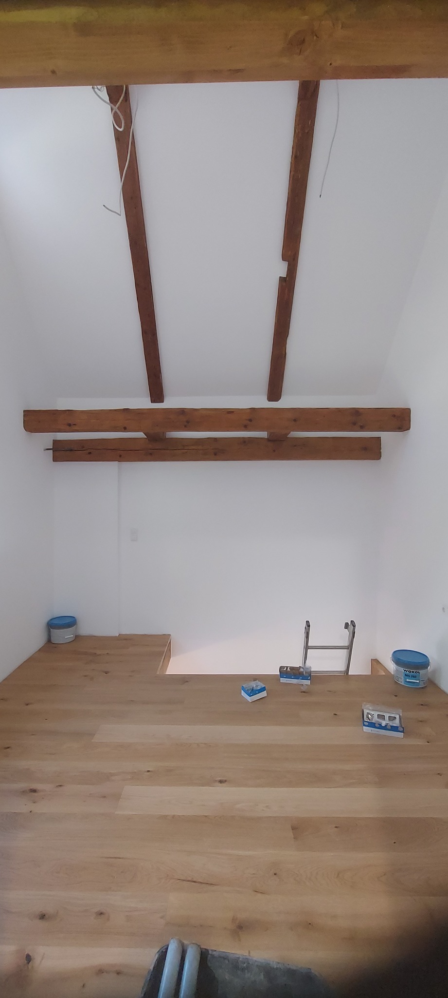 Jasna podłoga z paneli drewnianych w pokoju z drewnianymi belkami stropowymi. Widoczne akcesoria montażowe i drabina. Świeżo wykończone wnętrze.