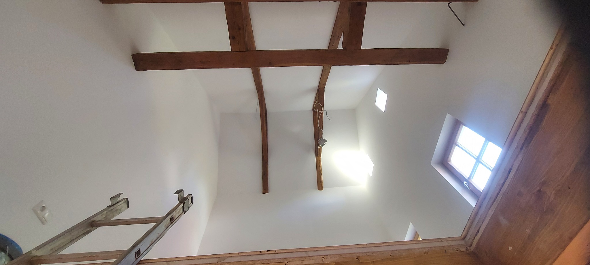 Białe wnętrze poddasza z drewnianymi belkami stropowymi, widok z dołu. Fragment drabiny malarskiej w lewym dolnym rogu. Okno dachowe w prawym górnym rogu.