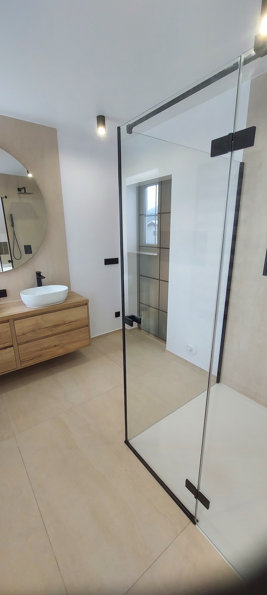 Nowoczesna łazienka z kabiną prysznicową typu walk-in, umywalką nablatową na drewnianej szafce i okrągłym lustrem. Beżowe płytki podłogowe i ściany w neutralnych odcieniach.