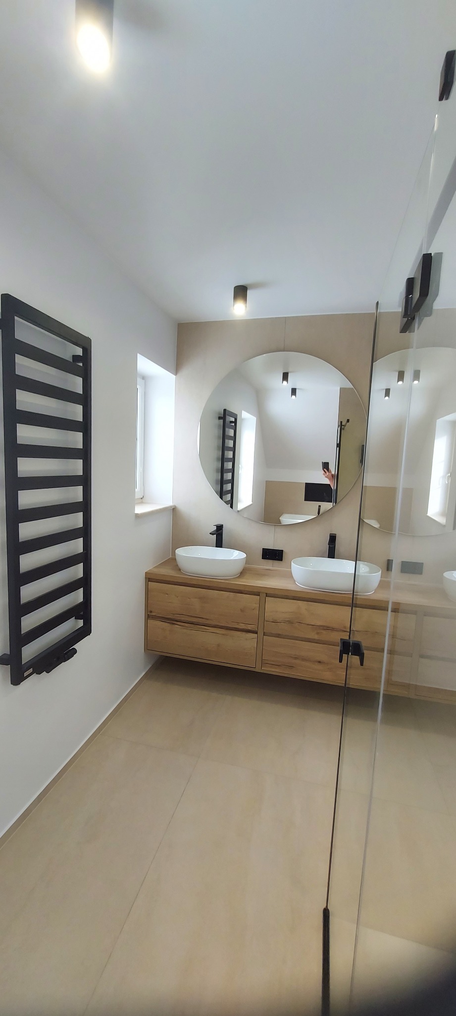 Nowoczesna łazienka z drewnianą szafką pod umywalki, okrągłym lustrem i czarnym grzejnikiem drabinkowym. Beżowe płytki podłogowe i kabina prysznicowa ze szkła.