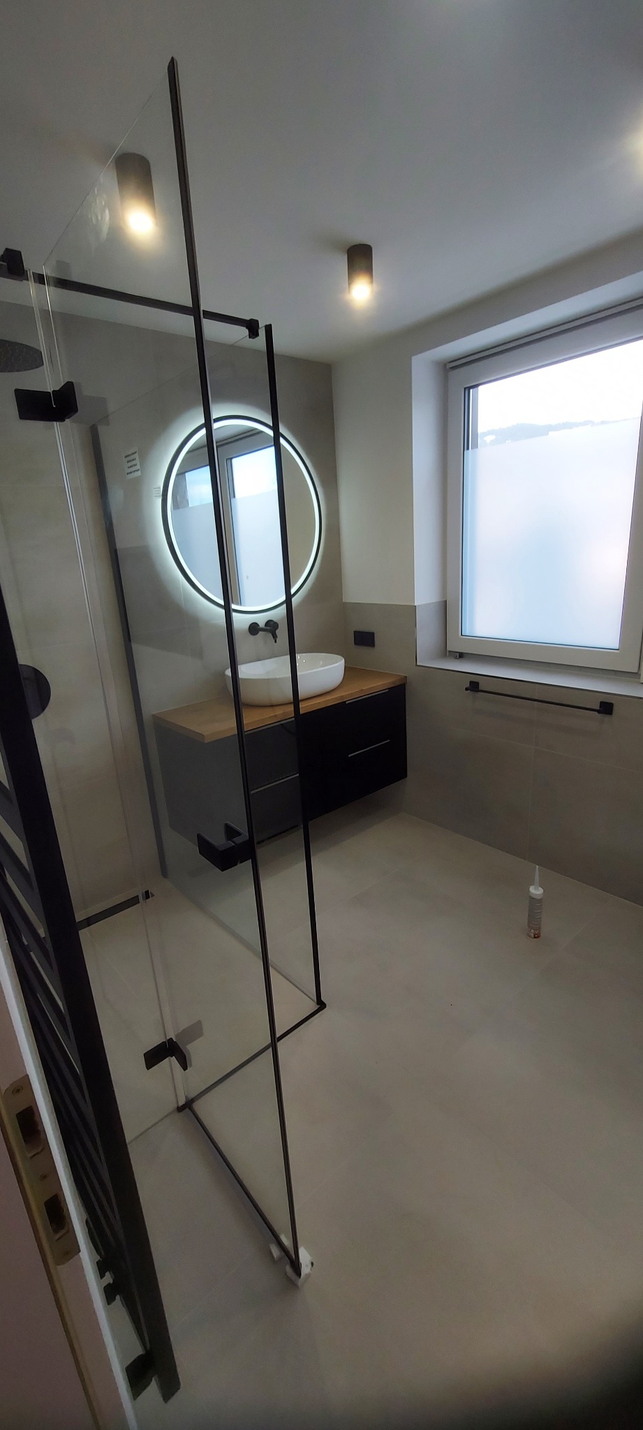 Nowoczesna łazienka z kabiną prysznicową typu walk-in, okrągłym lustrem z podświetleniem LED i minimalistyczną umywalką nablatową. Jasne płytki i czarne akcenty.