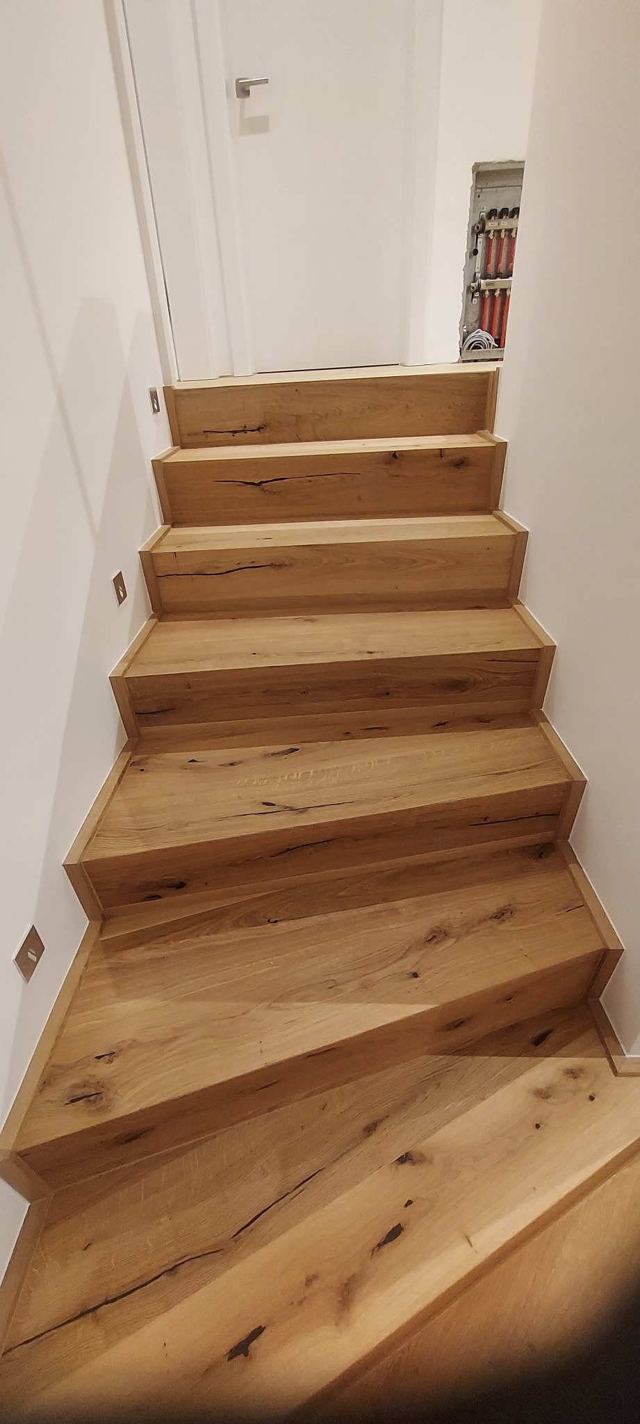 Drewniane schody z oświetleniem punktowym wzdłuż białej ściany. Widoczne sęki i naturalne usłojenie drewna. Ujęcie z dołu, podkreślające perspektywę.