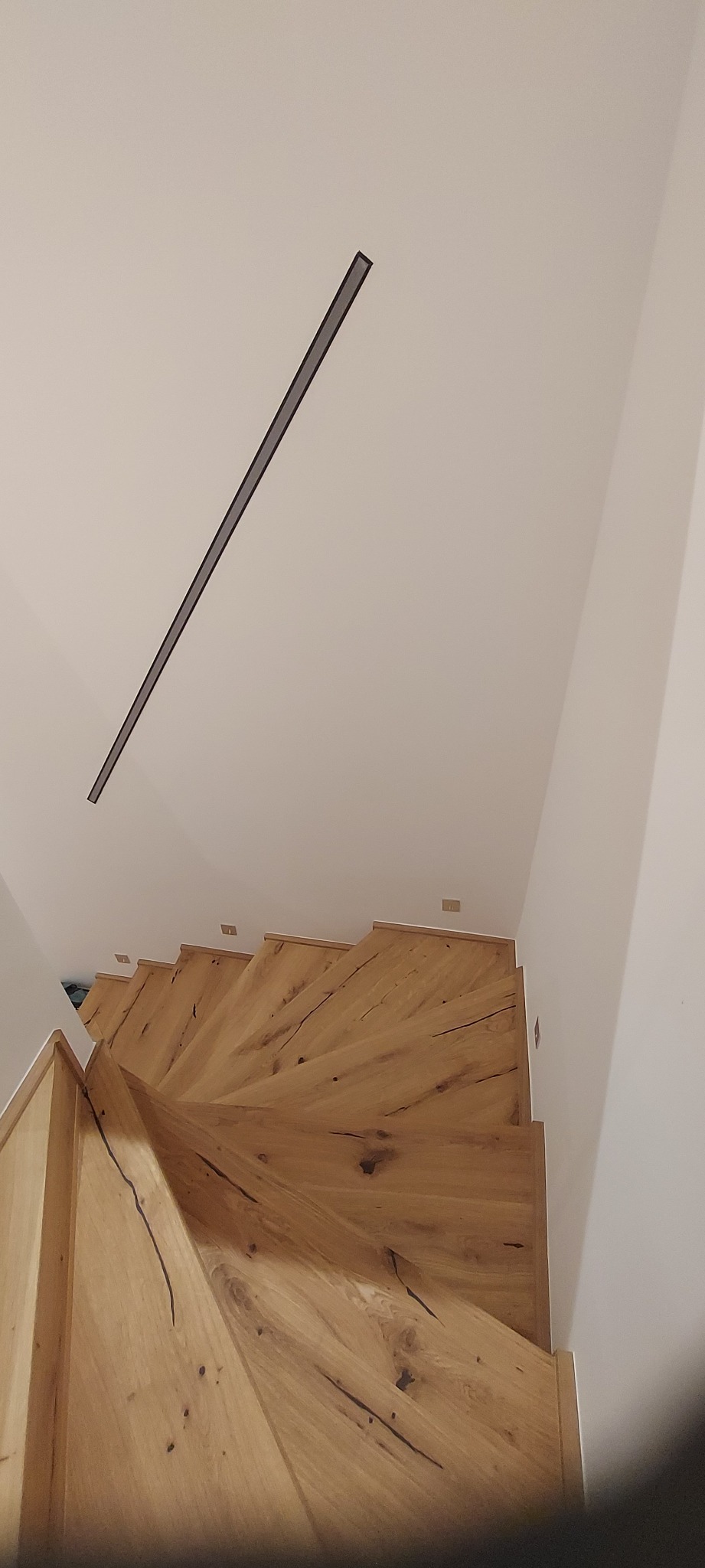 Drewniane schody z widocznymi słojami drewna, oświetlone listwą LED na suficie. Ujęcie z góry podkreśla spiralny kształt schodów i naturalną teksturę drewna.